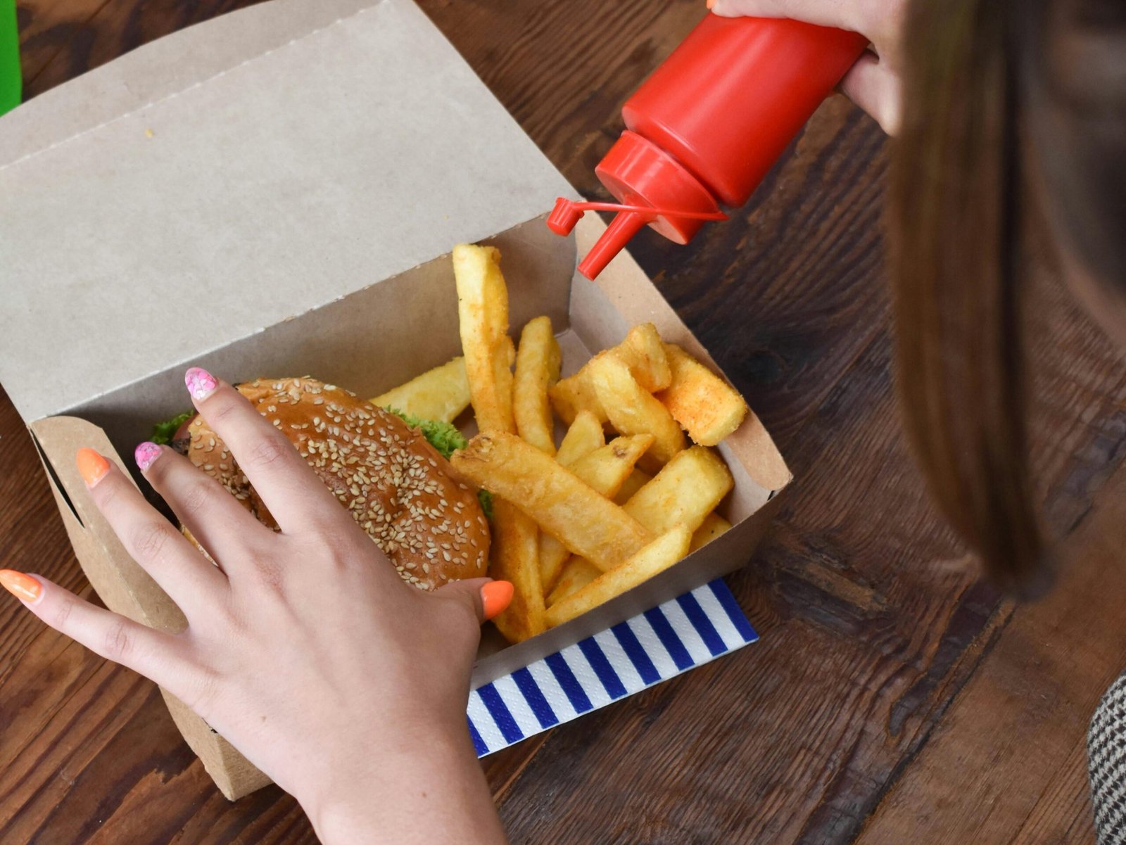 Overhead view of a woman adding ketchup to a burger and fries combo in a paper box.