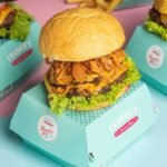 Close-up of a gourmet burger with onion rings and lettuce, served in a turquoise box.