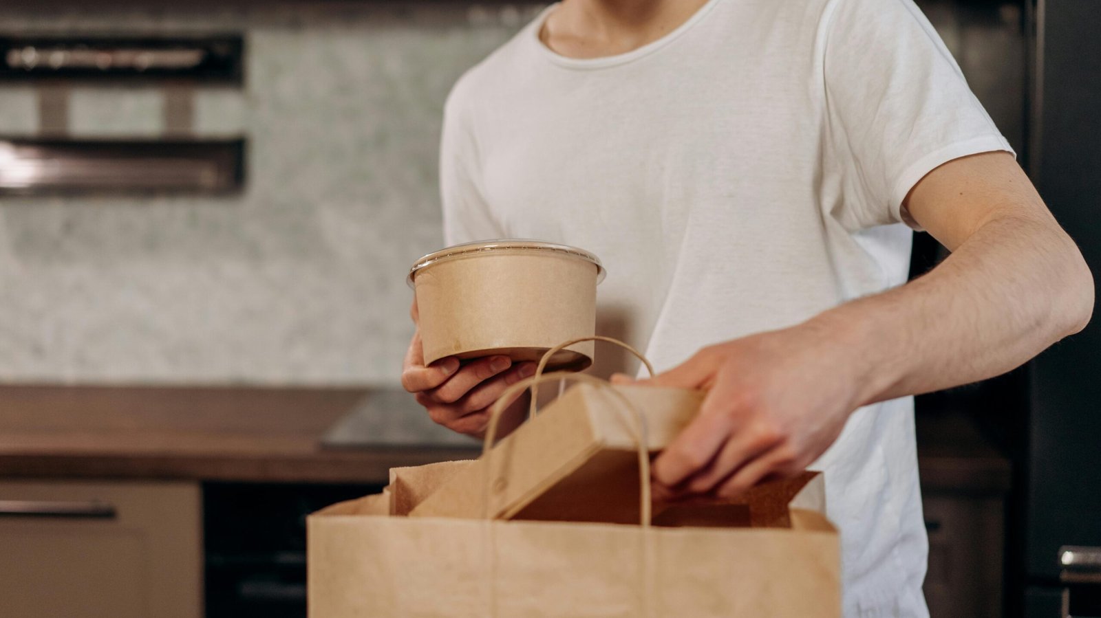 Man placing takeout containers into paper bag in kitchen setting.