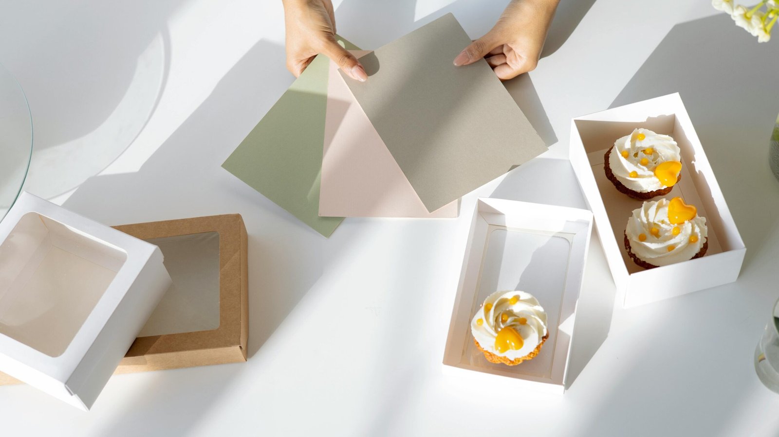 Hands holding color swatches over cupcakes and packaging in a bright setting.