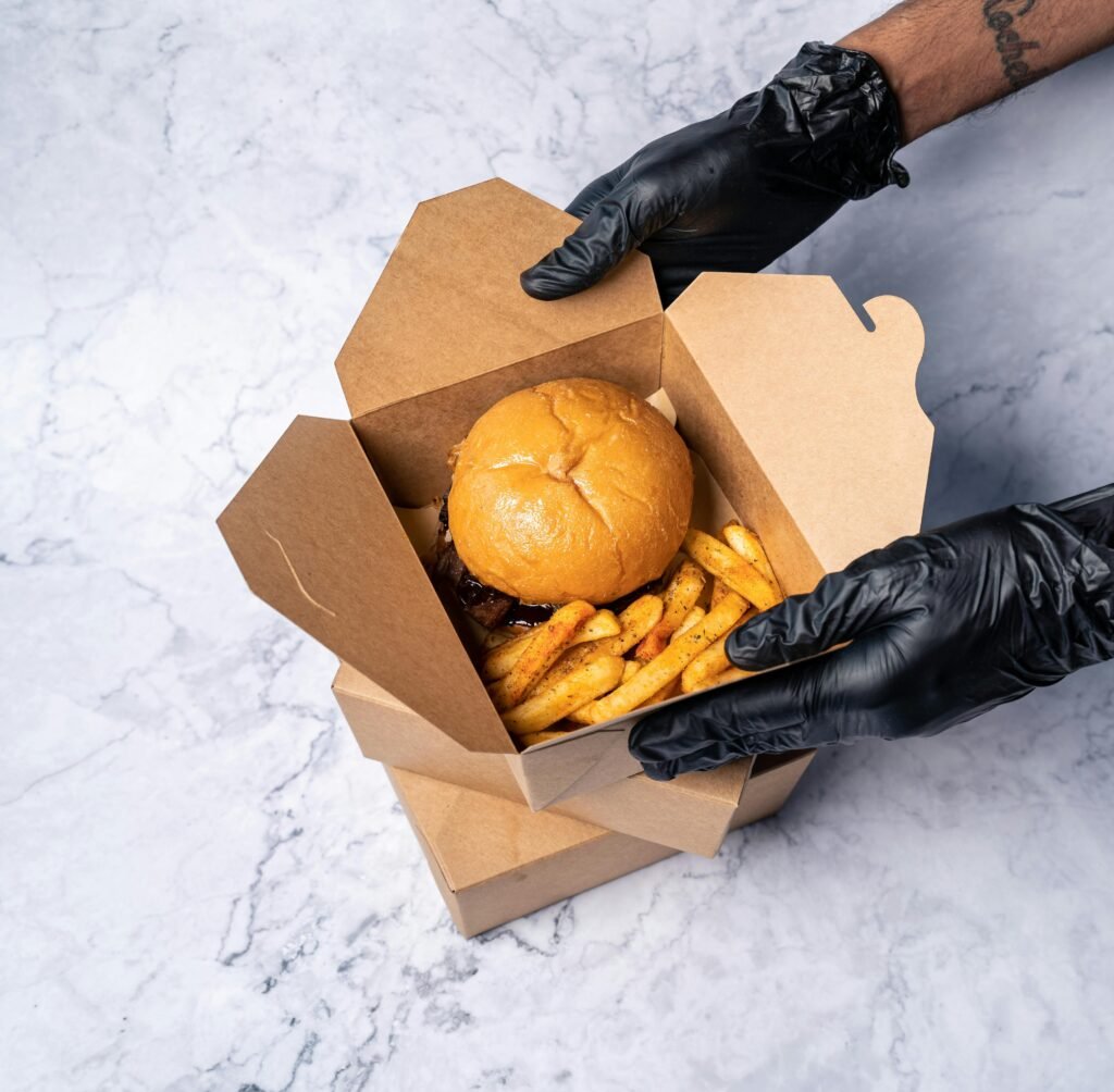 Delicious burger and seasoned fries in a cardboard takeout box, held by gloved hands.