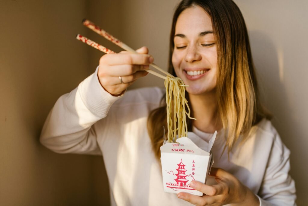 Smiling woman relishing Asian noodles using chopsticks in a cozy indoor setting.