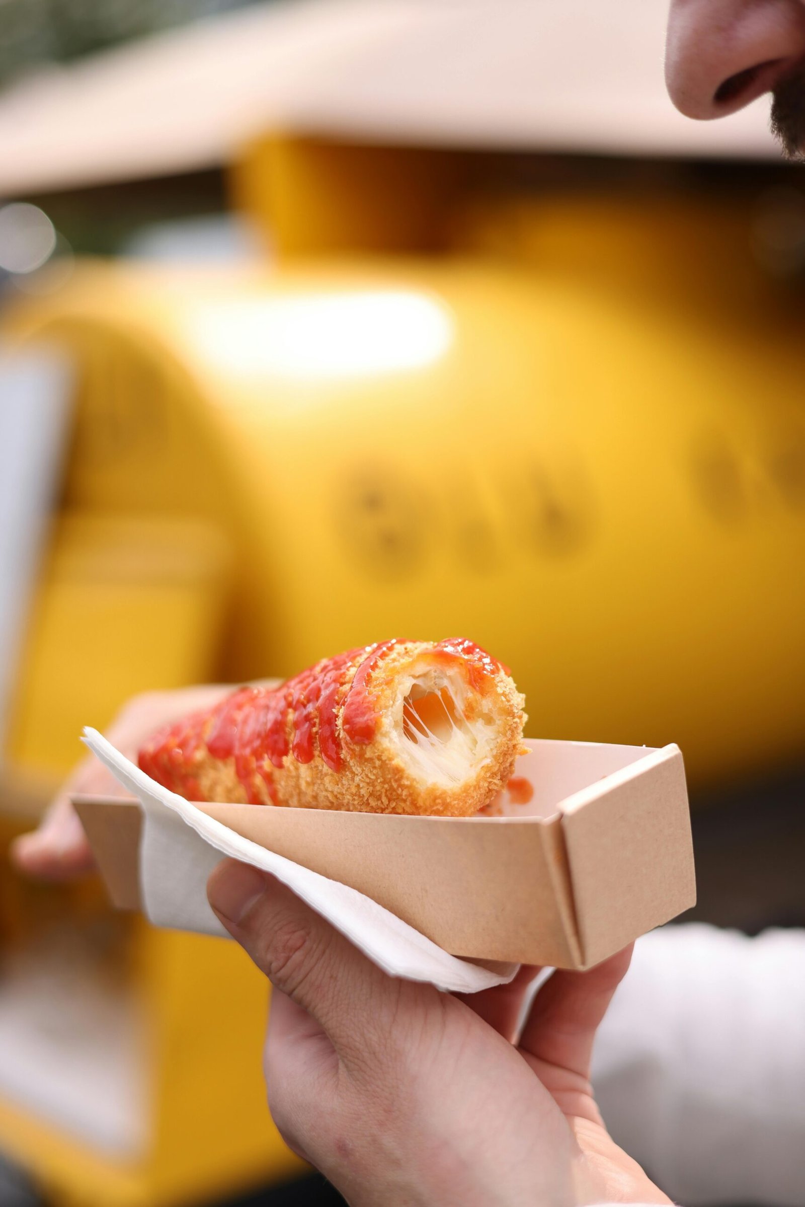 Close-up of a man holding a crispy cheese corn dog topped with ketchup, outdoors.