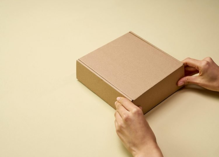 A close-up of hands opening a plain brown cardboard box on a beige surface, perfect for themes of shipping or packaging.